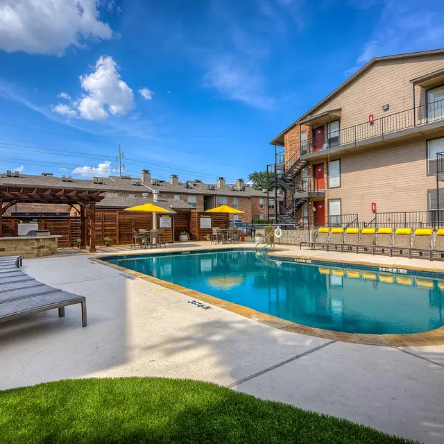 A swimming pool area with lounge chairs, surrounded by green grass and an apartment building in the background. There are umbrellas providing shade and a clear blue sky with scattered clouds overhead.