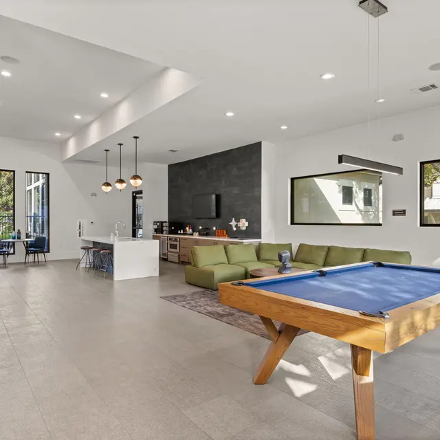 A spacious game room featuring a billiard table, a green sectional sofa, and large windows that let in natural light. The room has a modern design with a neutral color palette, and includes seating areas and pendant lighting.