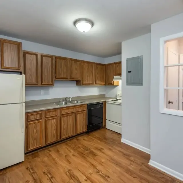 A modern kitchen featuring wooden cabinets, a white refrigerator, and a range and oven. The floor is wooden and the walls are painted light gray.