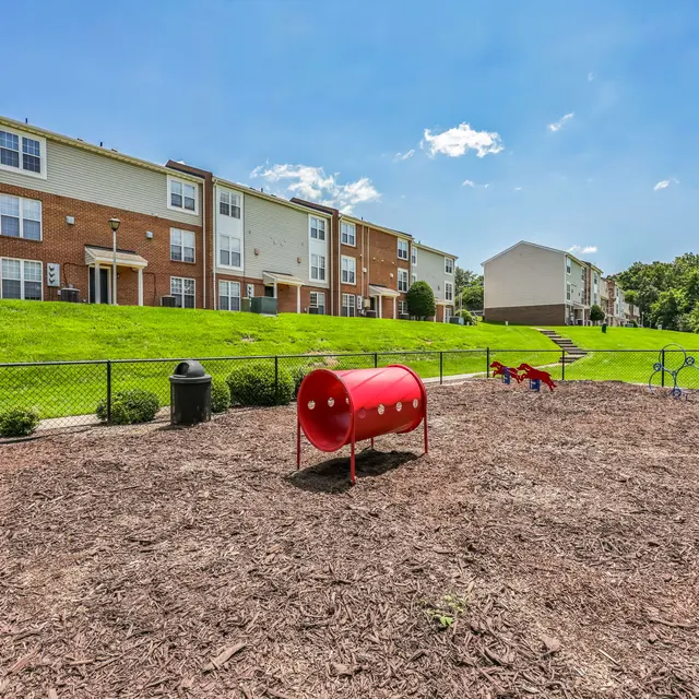 Playground in a Residential Community A playground area featuring a red tube slide and mulch ground cover, surrounded by a fenced area with seating and apartment buildings in the background.