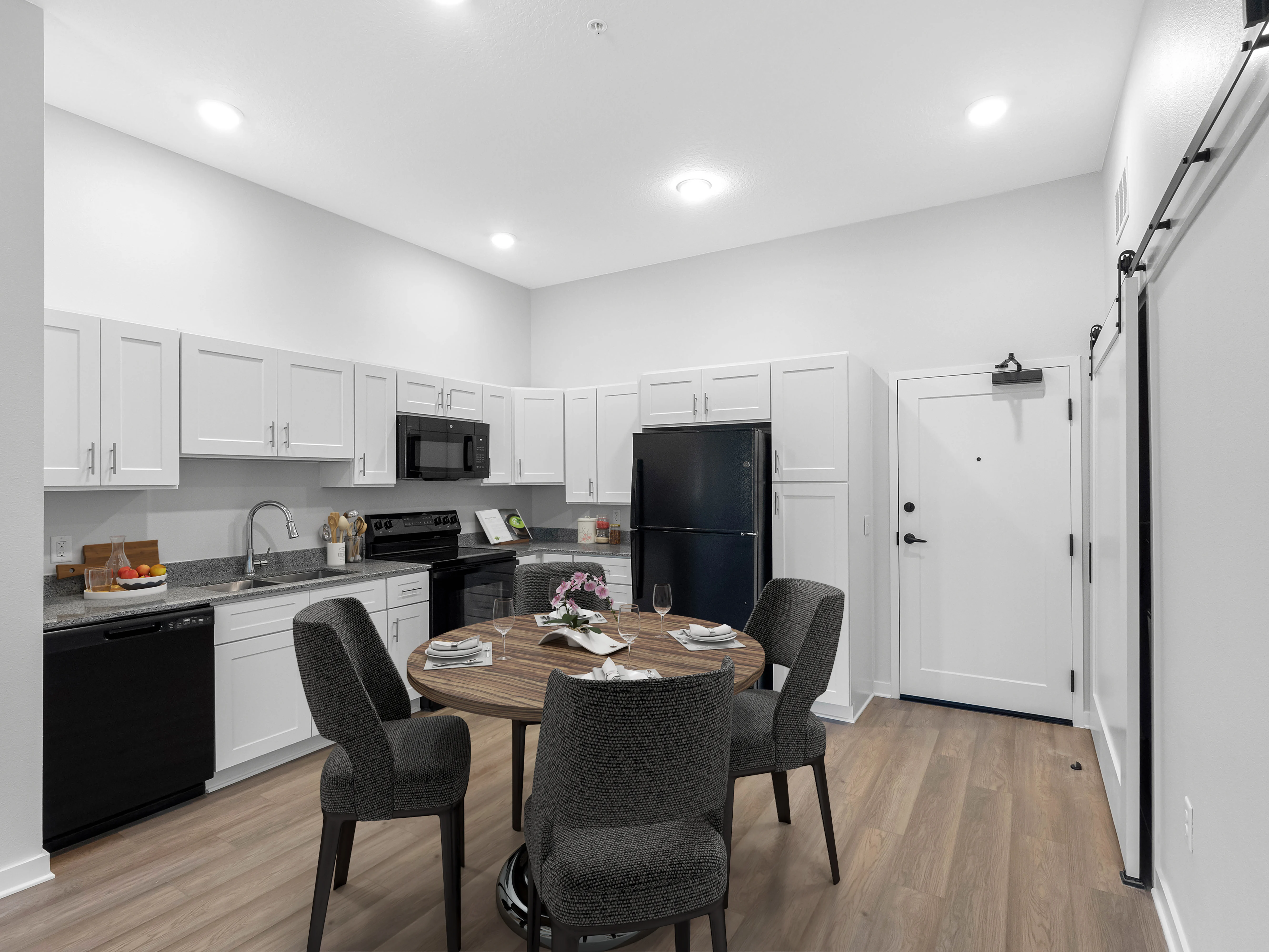 A modern kitchen featuring white cabinetry, a black refrigerator, a black dishwasher, and a circular wooden dining table with four chairs. The space is well-lit with recessed lighting and has a door leading outside.