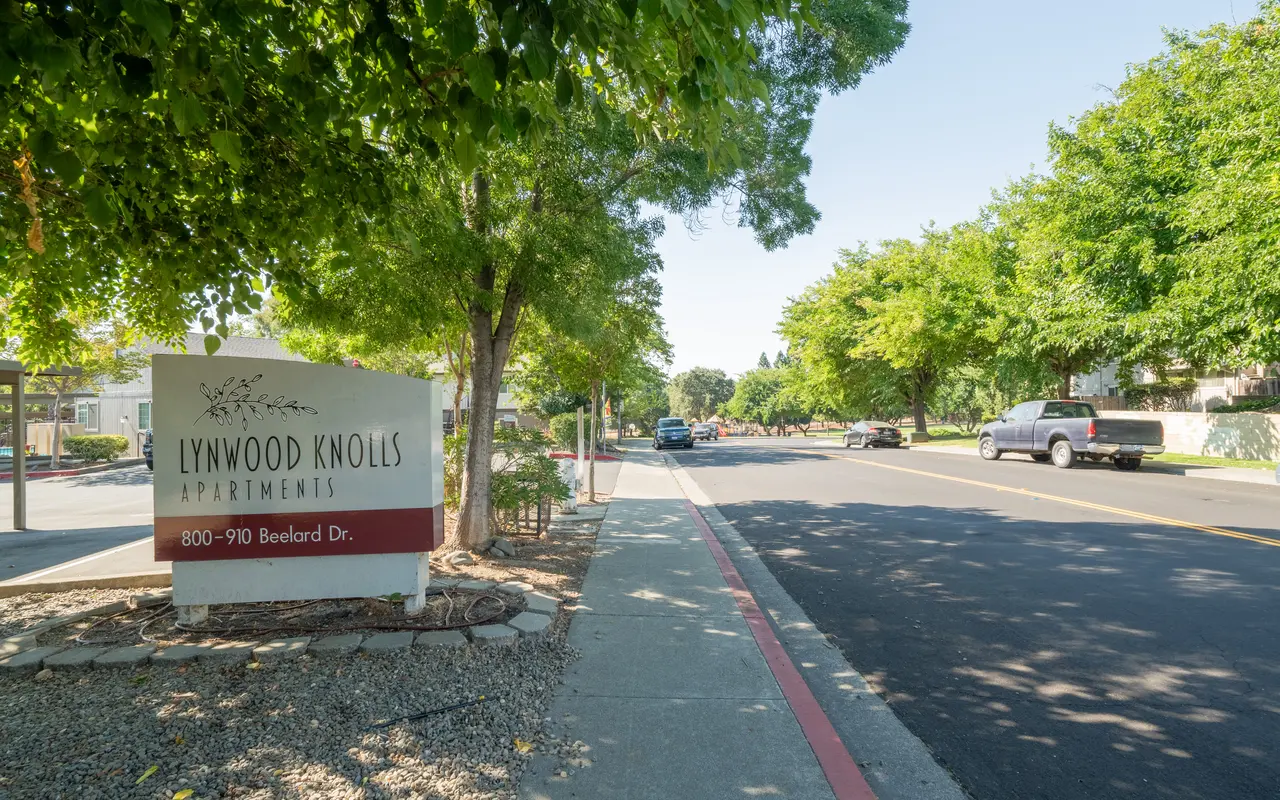 Entrance to Lynwood Knolls Apartments with trees lining the street on a sunny day.