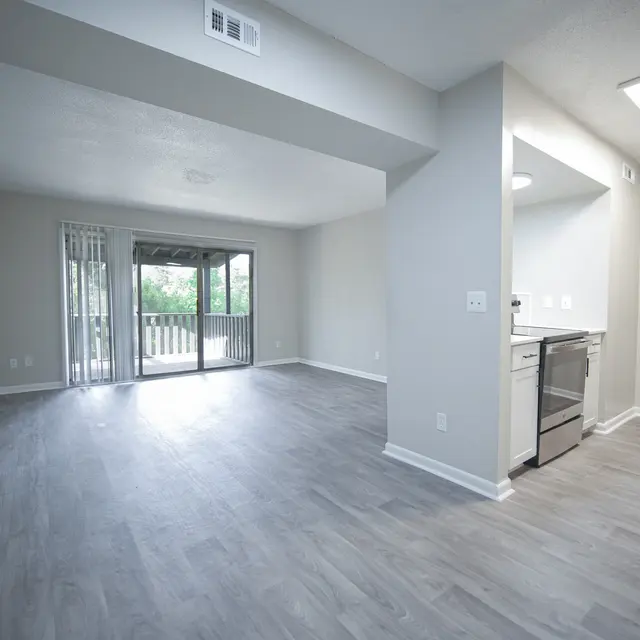 A spacious, modern apartment interior featuring an open living area and kitchen. Large windows allow natural light to flow in, showcasing a light gray color scheme and sleek flooring.