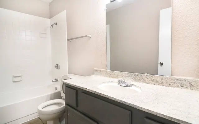 A clean and modern bathroom featuring a white bathtub, toilet, and a granite counter with a sink. The walls are painted in a light color, creating a bright and airy atmosphere. A large mirror is mounted above the sink, and a towel rack is visible on the wall.