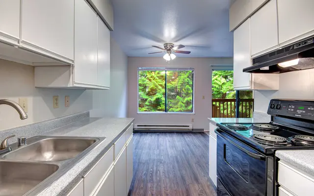 A modern kitchen with white cabinets and appliances, featuring a double sink and a black stove. A large window shows greenery outside, and the floor is dark wood.