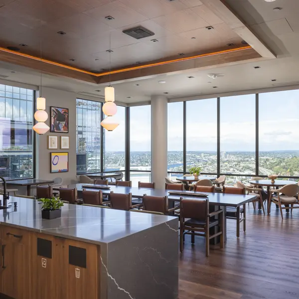 A spacious modern office lounge featuring large windows with a city view. The room is decorated with artwork on the walls, pendant lights hanging from the ceiling, and wooden tables and chairs arranged for casual seating. A kitchen counter with a plant is visible on the left side.