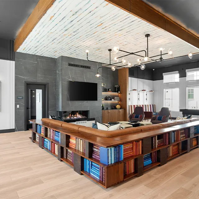 A modern lounge area featuring a stylish wooden sofa with colorful books as the base, a large flat-screen TV, and elegant lighting. There is artwork on the wall and a cozy fireplace. The space is well-lit with large windows.