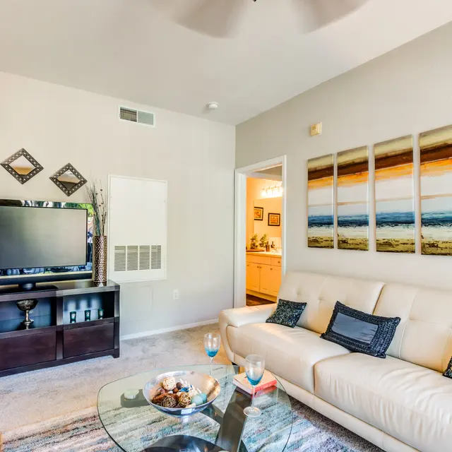 A modern living room featuring a white leather sofa, a glass coffee table, and a large television on a dark wooden console. The wall displays a sequence of landscape paintings with warm colors. Decorative elements include plants and geometric mirrors.