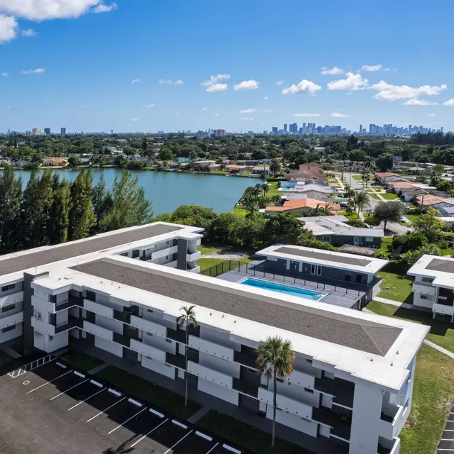 An aerial view of a modern apartment complex next to a lake, with a swimming pool and surrounding residential houses.