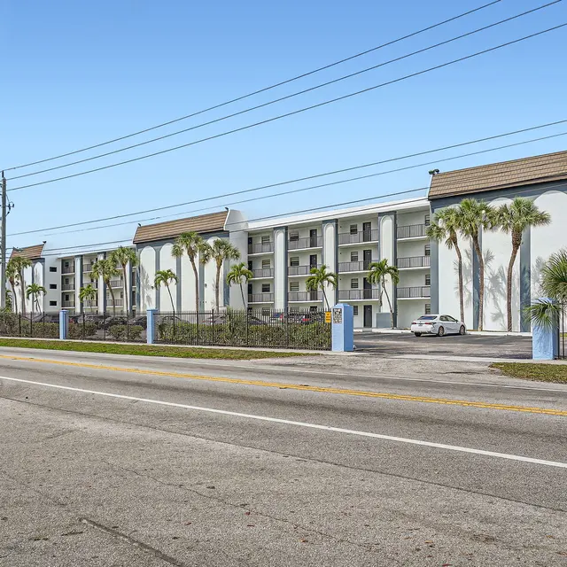 A modern apartment complex featuring multiple floors, palm trees, and a clear blue sky.