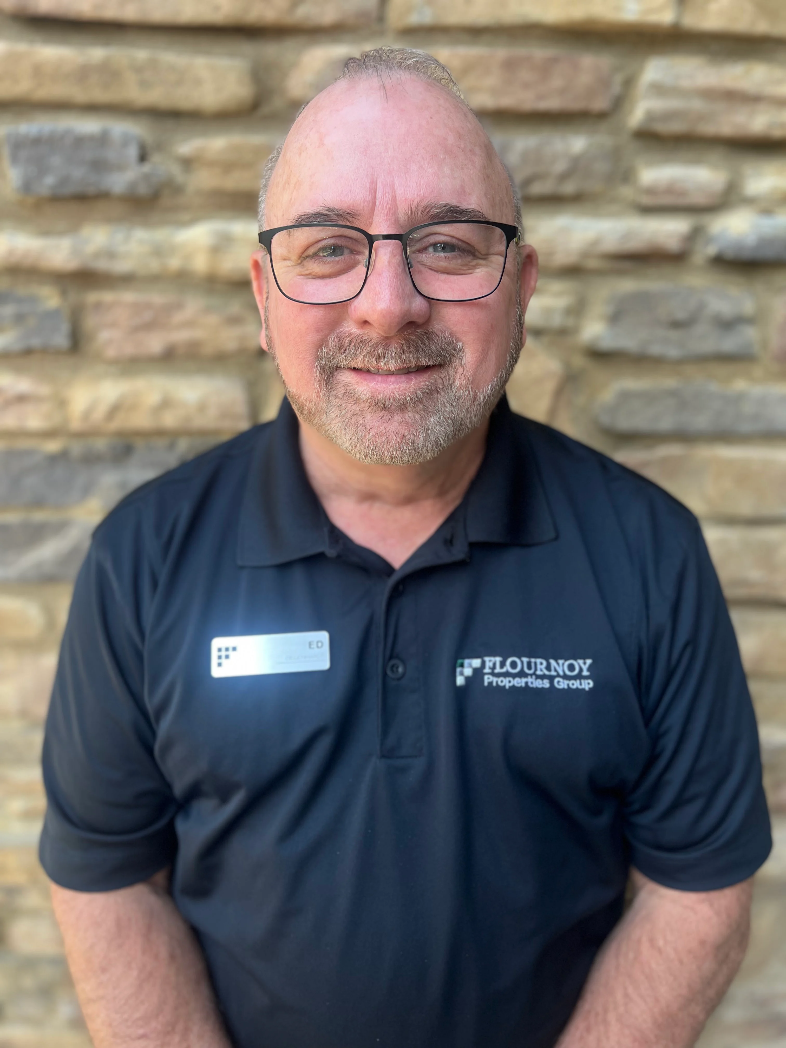 A smiling middle-aged man wearing glasses and a dark polo shirt with a name tag in front of a stone wall.