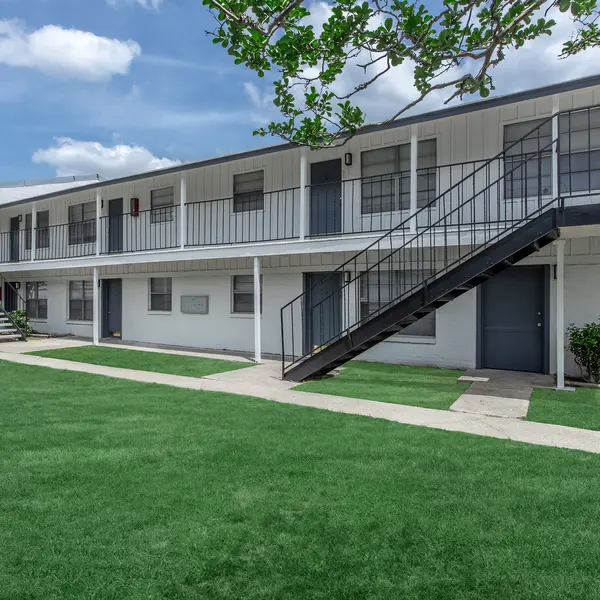 Exterior view of a two-story apartment complex with white walls, green grass, and a blue sky in the background.