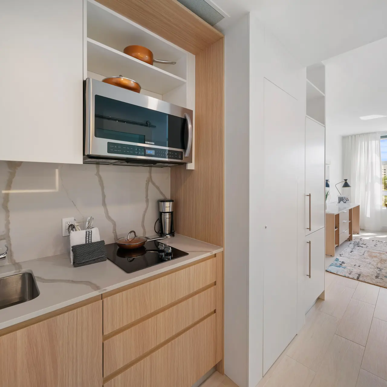 A modern kitchen interior with light wood cabinetry, a stainless steel sink, and a microwave above the counter. Natural light pours in from a nearby window into an adjoining living space with a table and chairs.