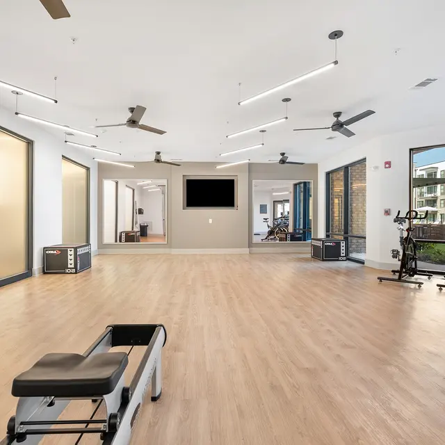 A spacious, bright gym interior featuring wooden flooring, workout machines, and large windows showing outdoor greenery.