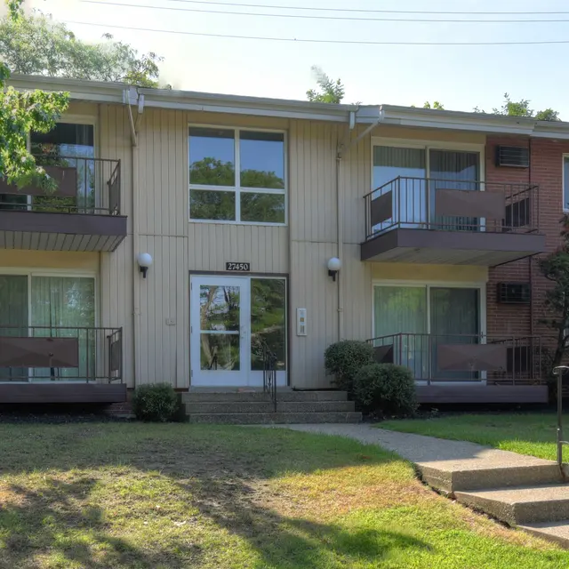 Front view of a two-story apartment building with balconies and large windows, surrounded by grass and trees.