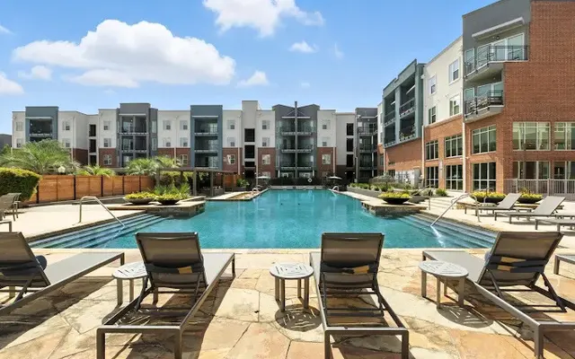 A tranquil pool area in a modern apartment complex, surrounded by lounge chairs and patio spaces, with well-maintained landscaping and buildings in the background.