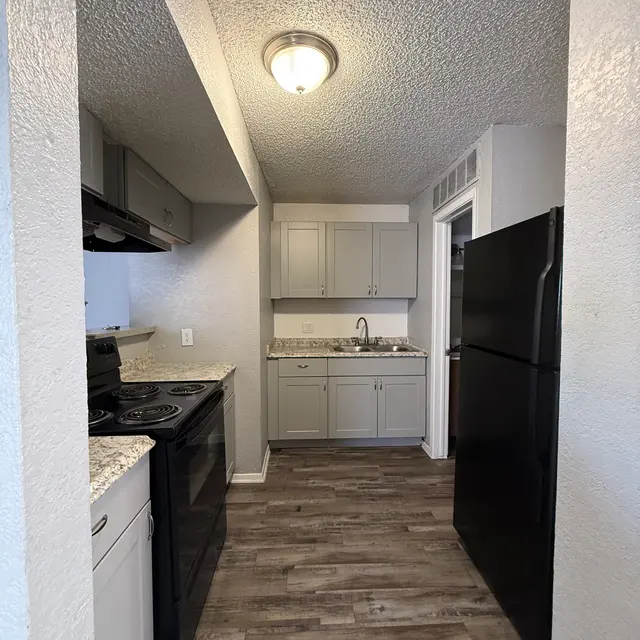 A modern kitchen featuring white cabinets and a black refrigerator. The kitchen has a granite countertop with a sink, an oven, and a stove. The floor is made of wooden planks, and the walls are painted in neutral tones. There's a ceiling light fixture.