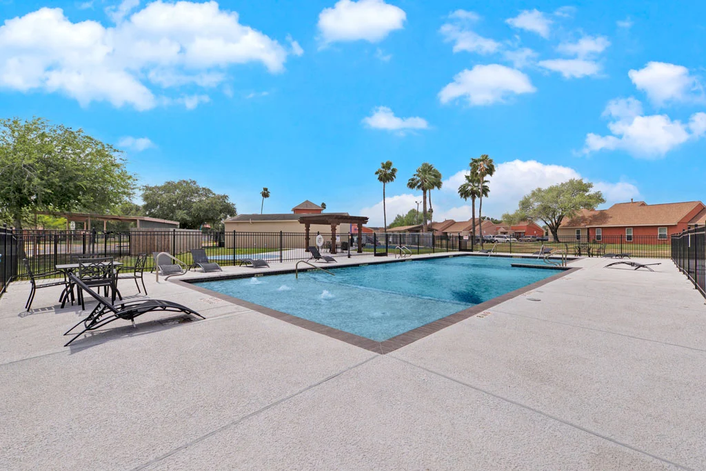 A clean, inviting swimming pool area surrounded by lounge chairs and palm trees, set against a blue sky with fluffy white clouds.