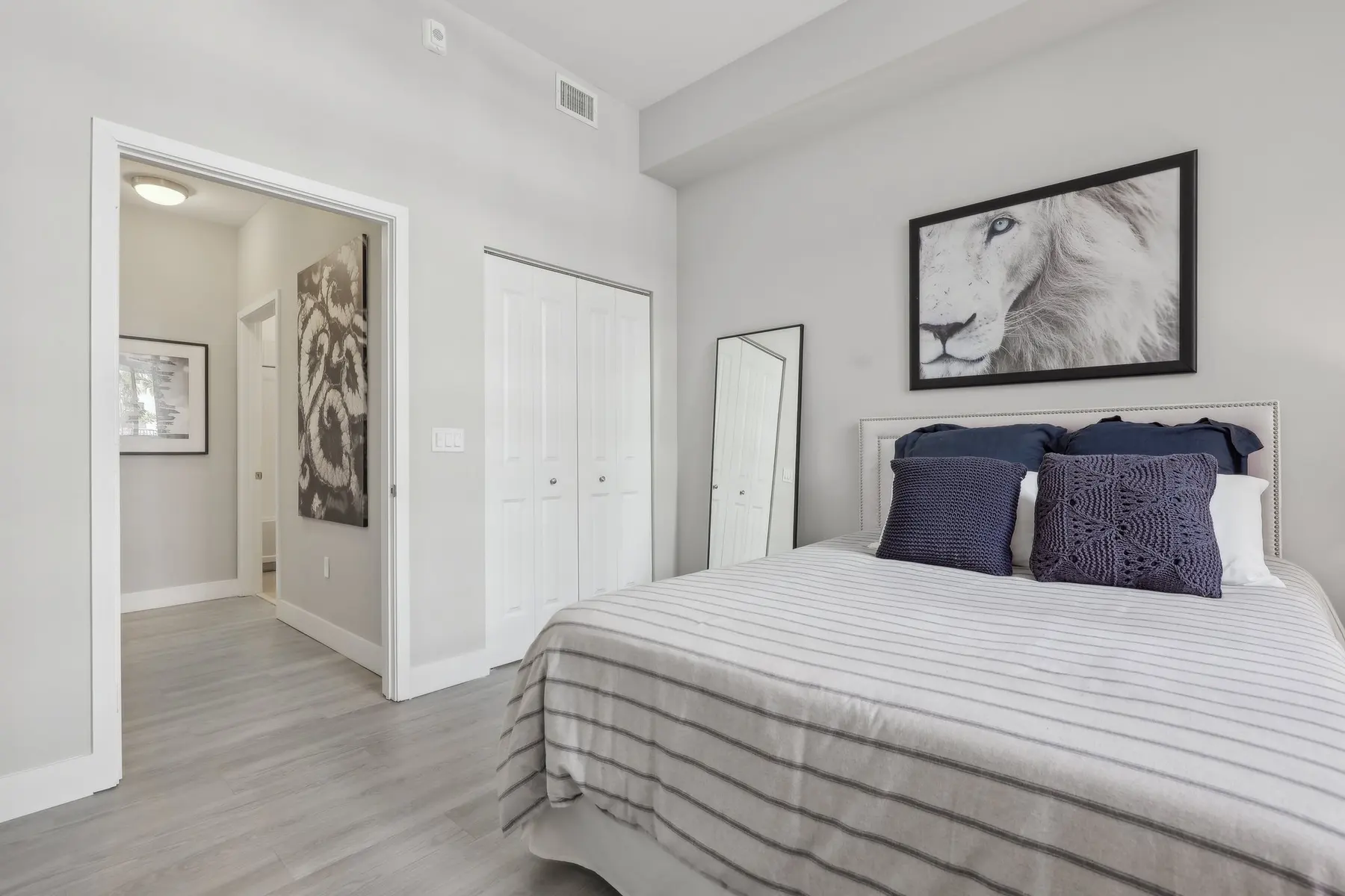 A modern bedroom featuring a bed with striped bedding, navy blue pillows, and a lion portrait on the wall. A full-length mirror stands next to the bed, and white closet doors are visible in the background along with a doorway leading to another room.