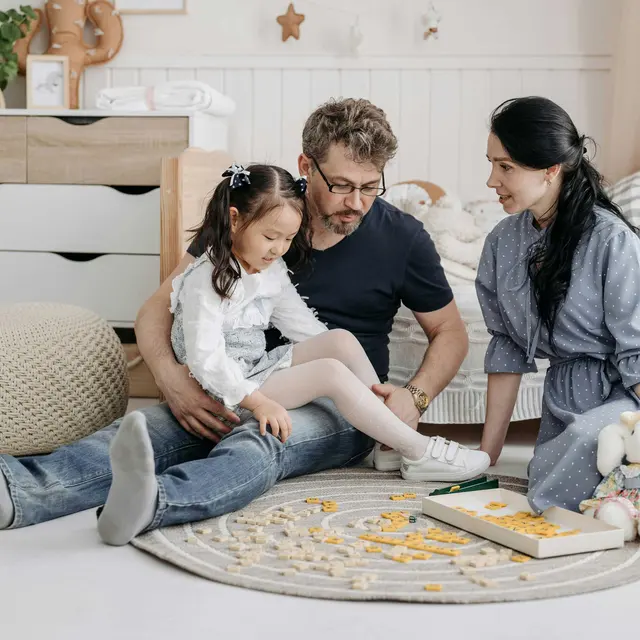Family Playing Together A family playing together on a rug in a cozy room. A child in a white outfit sits on the floor with her father, who wears a dark t-shirt and glasses. The mother, in a blue dress, engages with them. There are toys and a game on the rug.