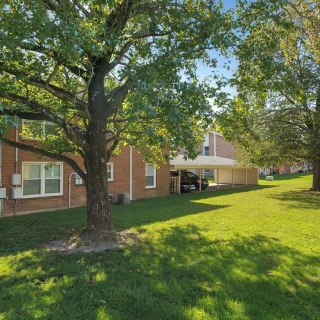 A peaceful park area featuring a large tree and well-maintained green lawn in front of a brick building with windows. A car is parked under a canopy nearby.