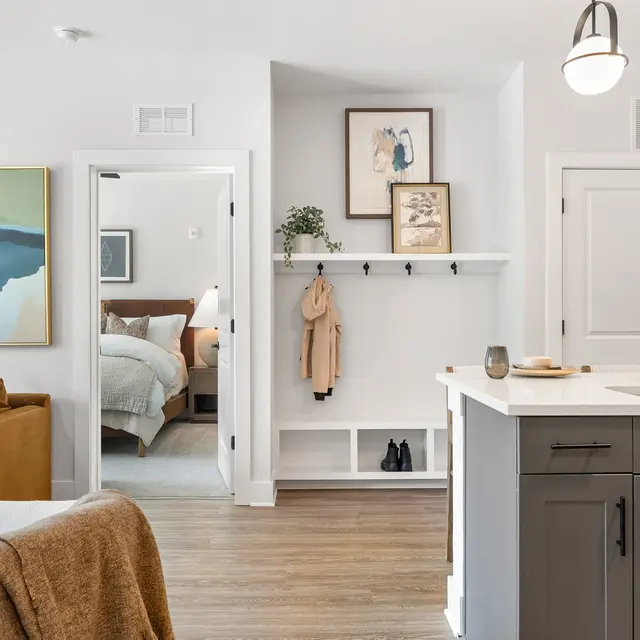 A modern apartment interior showing a kitchen area leading into a living room and a bedroom. The space has neutral colors with a cozy sofa, artworks on the wall, and hooks for hanging coats.