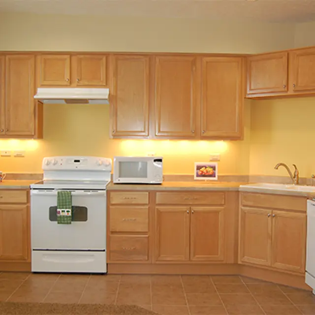 Modern Kitchen with Wooden Cabinets A well-lit modern kitchen featuring wooden cabinets, a white stove, a microwave, and a refrigerator. There is a green towel hanging on the stove and a small framed picture on the counter.