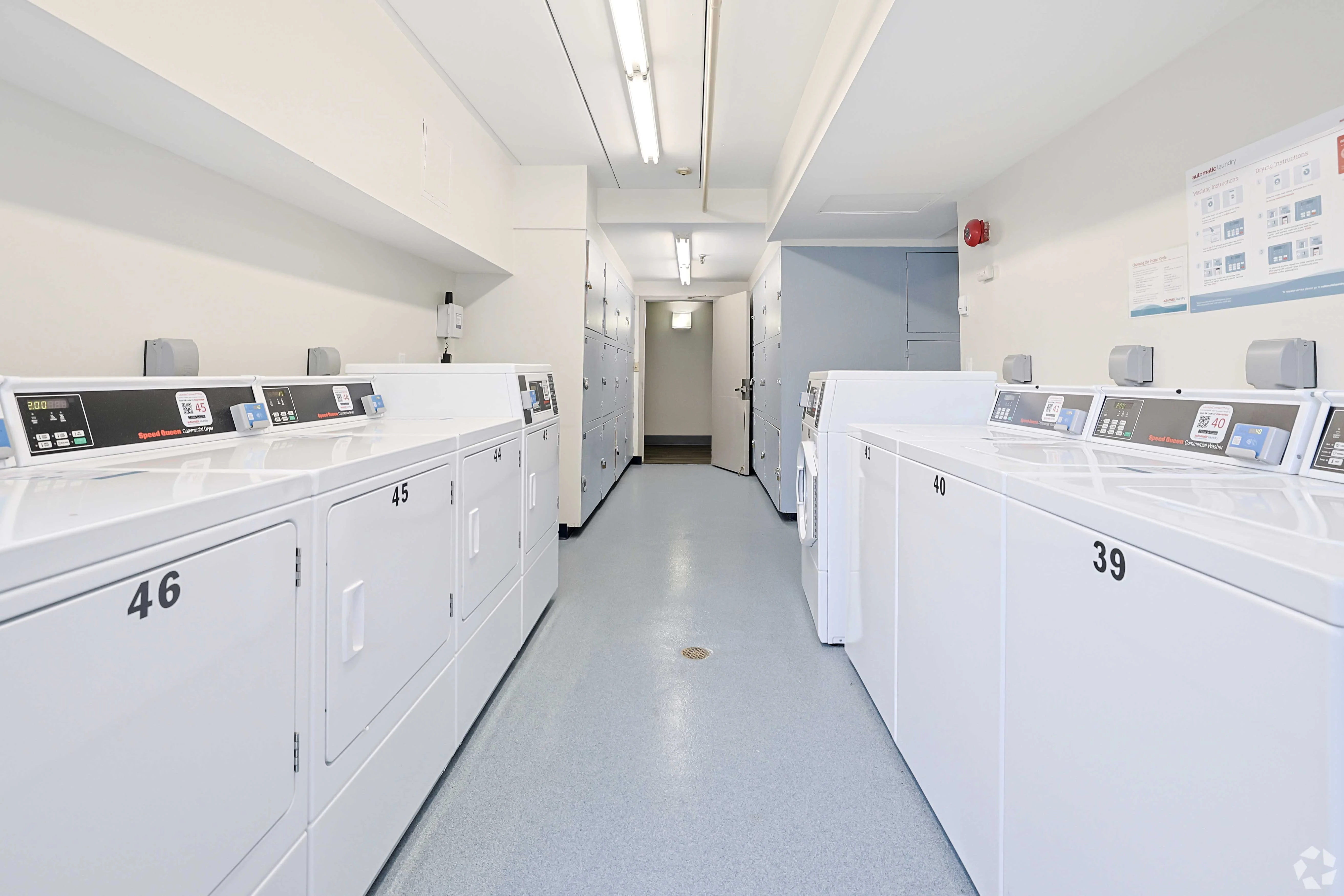A clean, modern laundry room featuring several stacked washing machines and dryers, numbered 39, 45, and 46, arranged in rows. The space is well-lit with a light grey floor and white walls, leading to a door at the end.