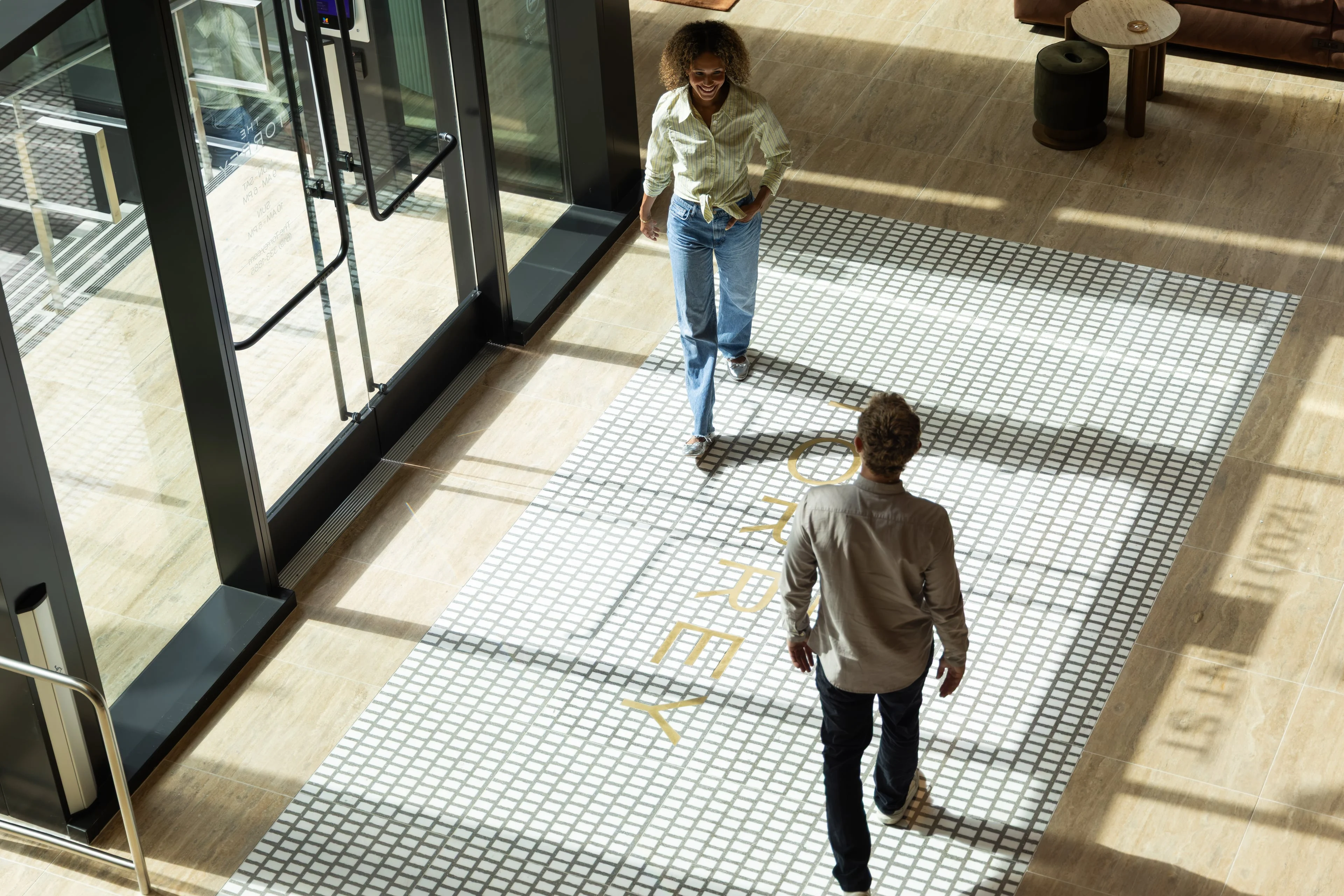 Two individuals walking in a spacious, modern lobby with large windows and a tiled floor.
