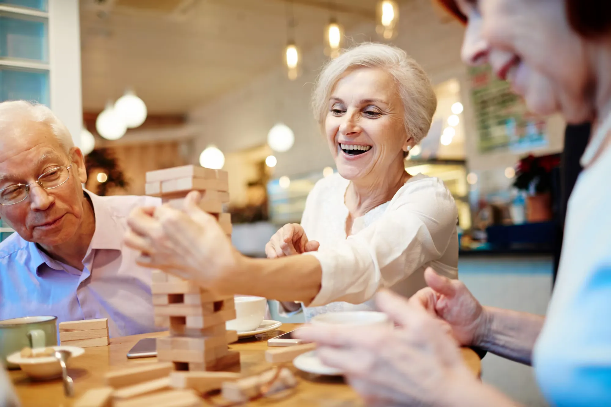 Joyful Moments of Seniors Playing a Game A group of seniors joyfully playing a game with wooden blocks at a cafe, with one woman reaching to add a block to a tower while the others watch.