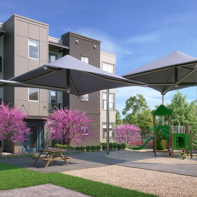 A modern playground area with green grass, picnic tables, and two large shaded structures, surrounded by blooming pink trees and an apartment building in the background.
