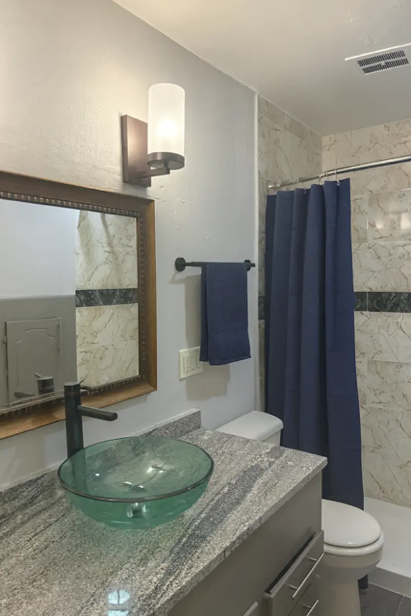 A modern bathroom featuring a round glass sink on a granite countertop, a large mirror with a frame, a towel rack with a blue towel, and a shower curtain in a coordinating blue color. Marble-like wall tiles add elegance to the space.