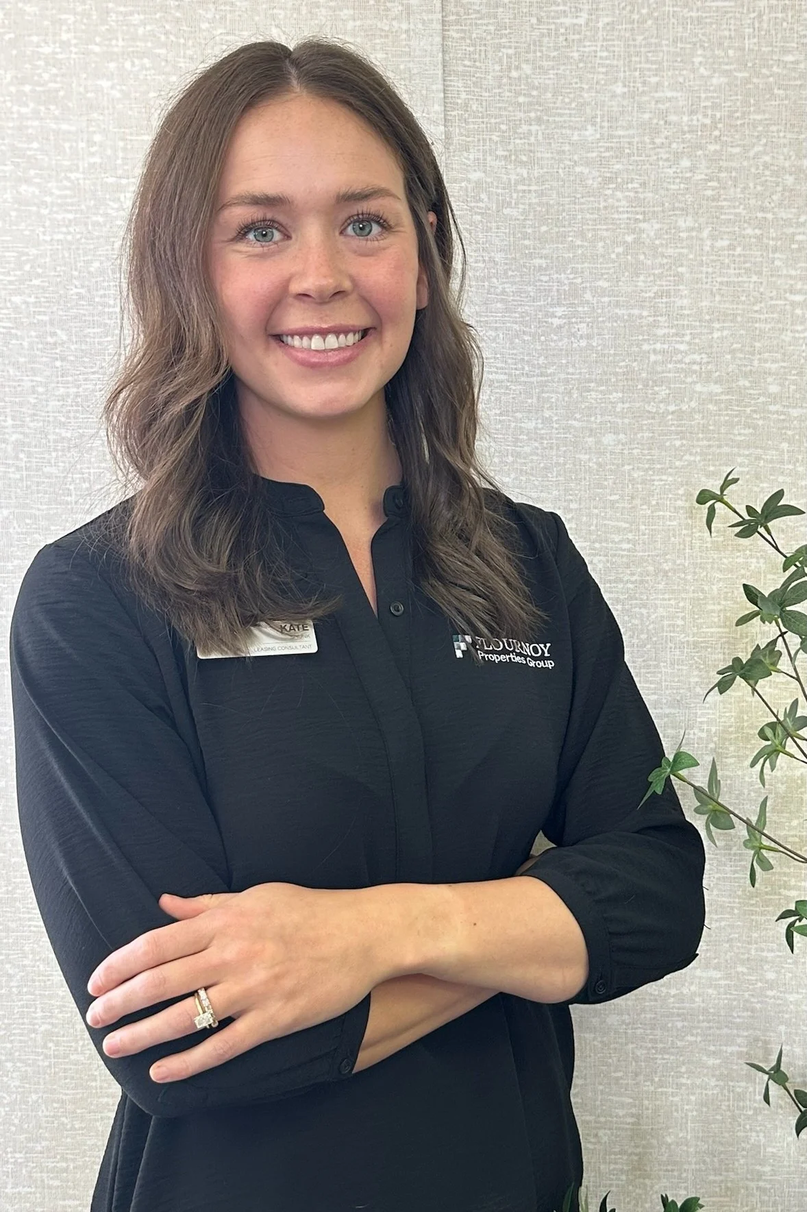 Professional Portrait of a Young Woman A woman with medium-length brown hair wearing a black shirt and a name tag, smiling while standing next to a plant.