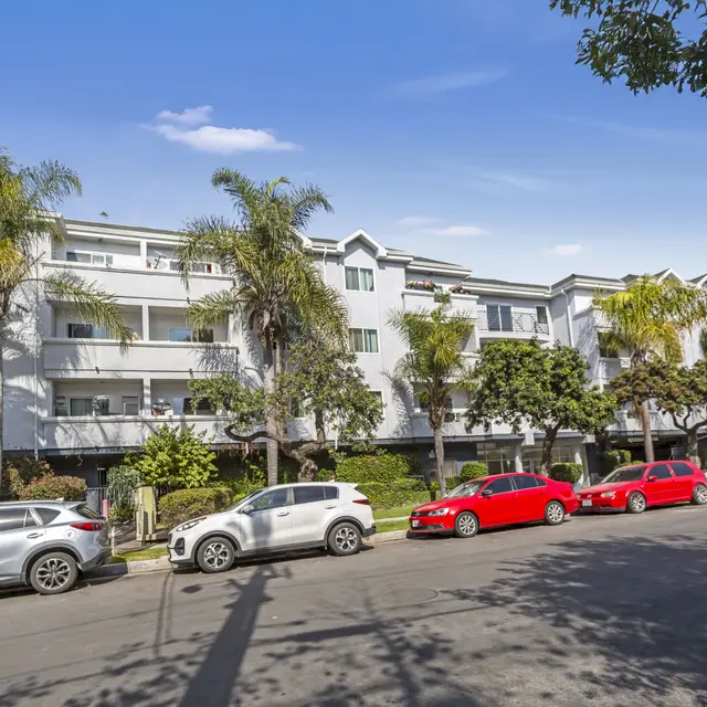 A street view of a three-story apartment building with palm trees and parked cars.
