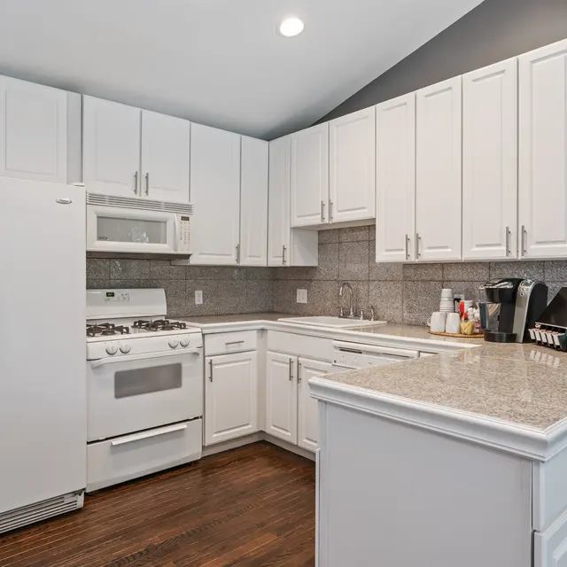 A modern kitchen featuring white cabinetry, a gas stove, and a large refrigerator. The countertops are a light stone color, and the room has hardwood flooring.