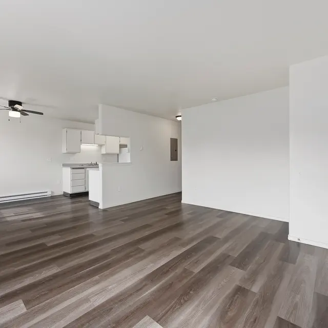 Spacious Modern Apartment Interior A spacious and modern apartment interior featuring wooden floors, a ceiling fan, and a kitchen area visible in the background. The walls are painted white, creating a bright and airy atmosphere.