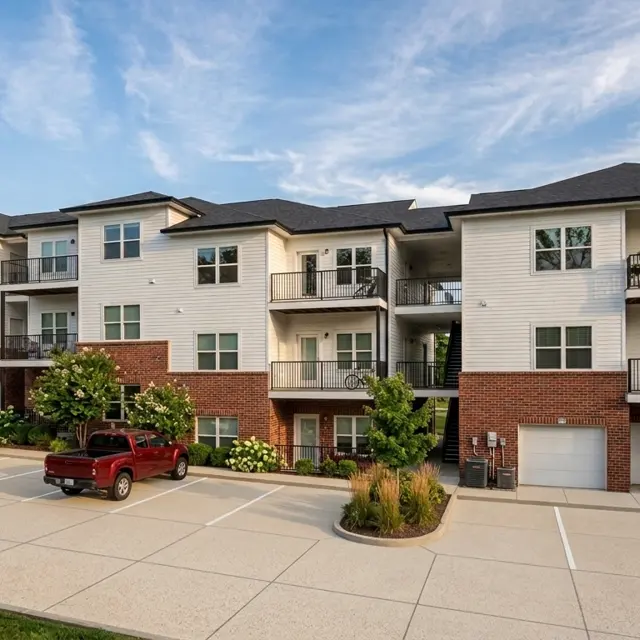 Exterior view of a modern apartment complex with multiple stories, surrounded by greenery and parked cars in the foreground.