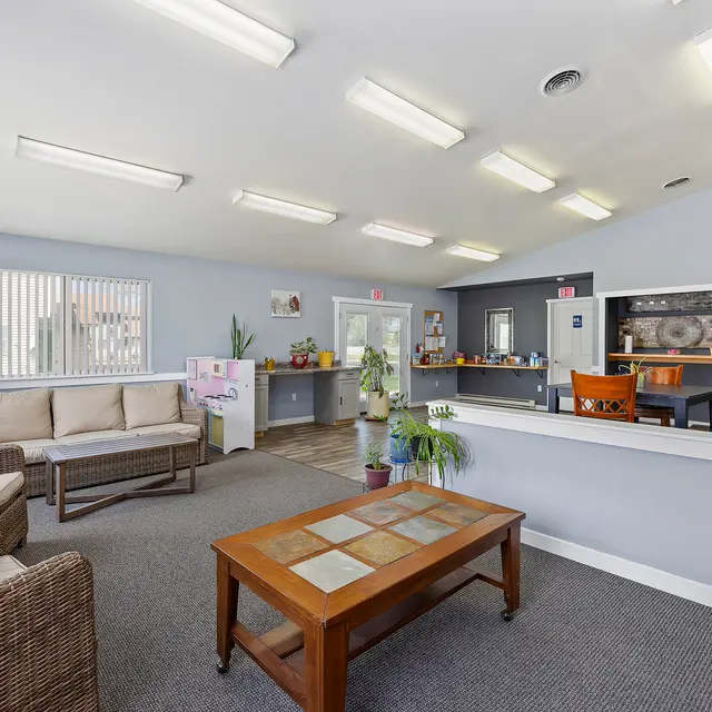 Spacious Office Lobby A spacious office lobby with comfortable seating and a reception area. The room features a large window with blinds, potted plants, and a wooden coffee table in the center.
