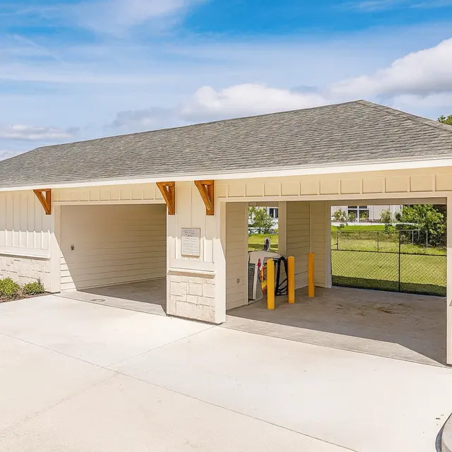 A spacious open carport structure with a sloped roof, featuring wooden beams and a concrete floor. The area is fenced and surrounded by greenery, with a stop sign visible nearby.