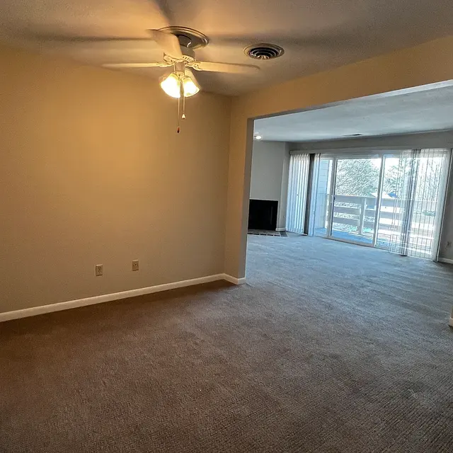 An open living space showing a room with carpet flooring, a ceiling fan, and a large window allowing natural light. In the background, there's another room with sliding glass doors leading to an outdoor area.