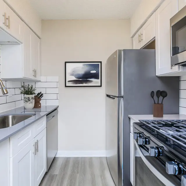 A modern kitchen featuring white cabinets, a stainless steel refrigerator, and a gas stove. The backsplash is made of white tiles, and there is a bowl of red apples on the counter.
