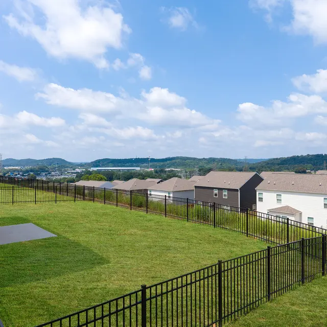 A panoramic view of a grassy backyard enclosed by a black fence, overlooking a neighborhood with houses and hills in the distance.
