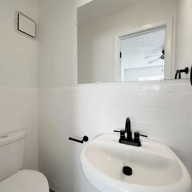 Small Modern Bathroom A small bathroom featuring a white tiled wall and a compact toilet next to a sink with a black faucet. A mirror is mounted above the sink, and a door leads to another room in the background.