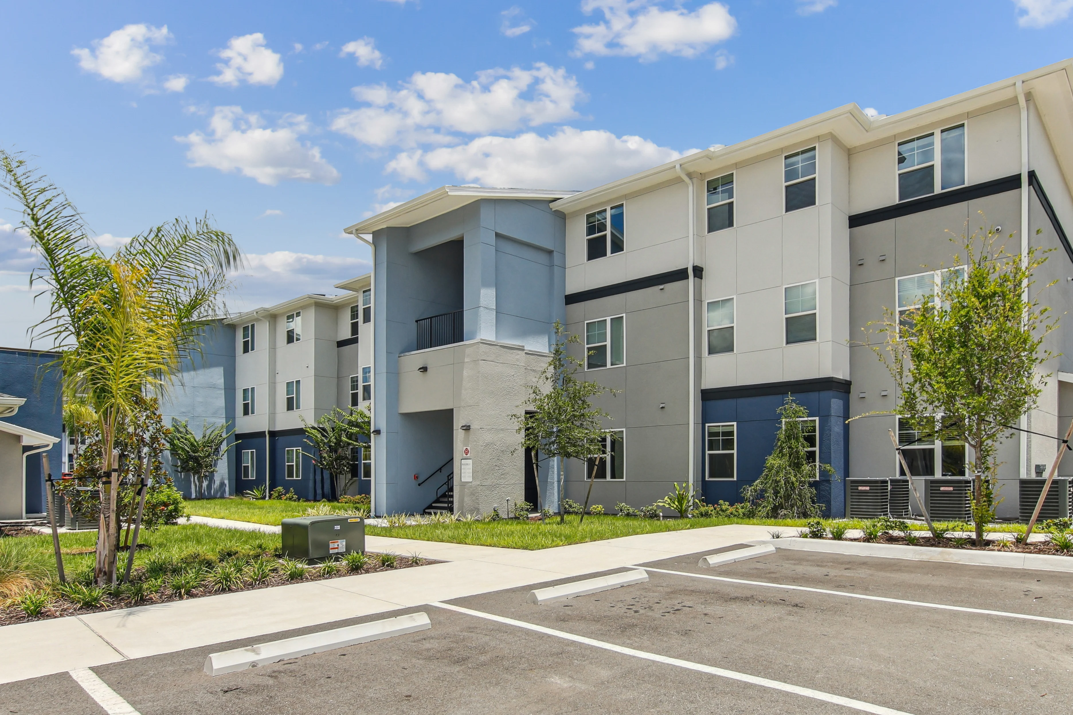 EKOS Lake Shadow A modern apartment complex with a three-story building, featuring a combination of blue and white siding, landscaped greenery, and a spacious parking area in front.