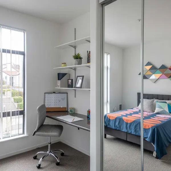 A modern bedroom featuring a grey bed with colorful bedding, a desk with a swivel chair, and large windows with blinds, showcasing a view of greenery outside.