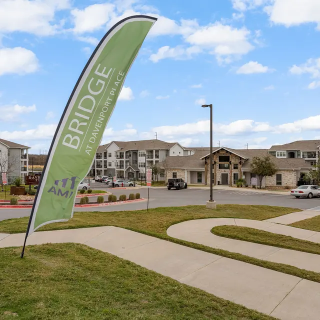 View of a residential apartment complex with a large flag displaying the word 'BRIDGE'. The scene includes roads, landscaped areas, and several buildings in the background under a partly cloudy sky.