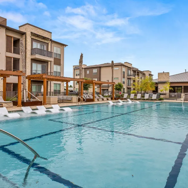 A modern apartment complex with a large swimming pool surrounded by lounge chairs and wooden pergolas on sunny weather.