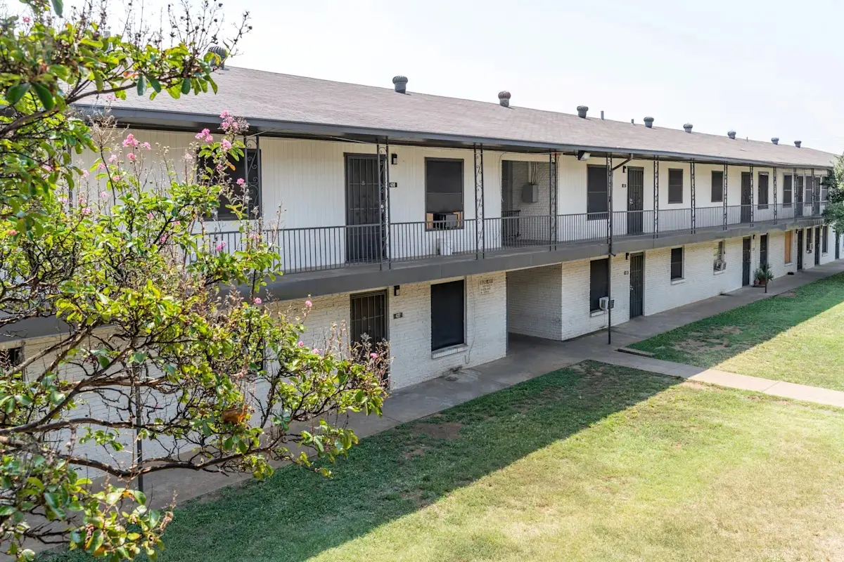 A view of a two-story apartment complex with a long porch and several doors, surrounded by grassy areas and a tree.