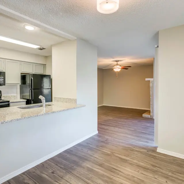 View of a modern kitchen with a granite countertop connected to a spacious living area. The kitchen has a black stove and microwave, along with a black refrigerator. The living area features a ceiling fan and has wood-like flooring.