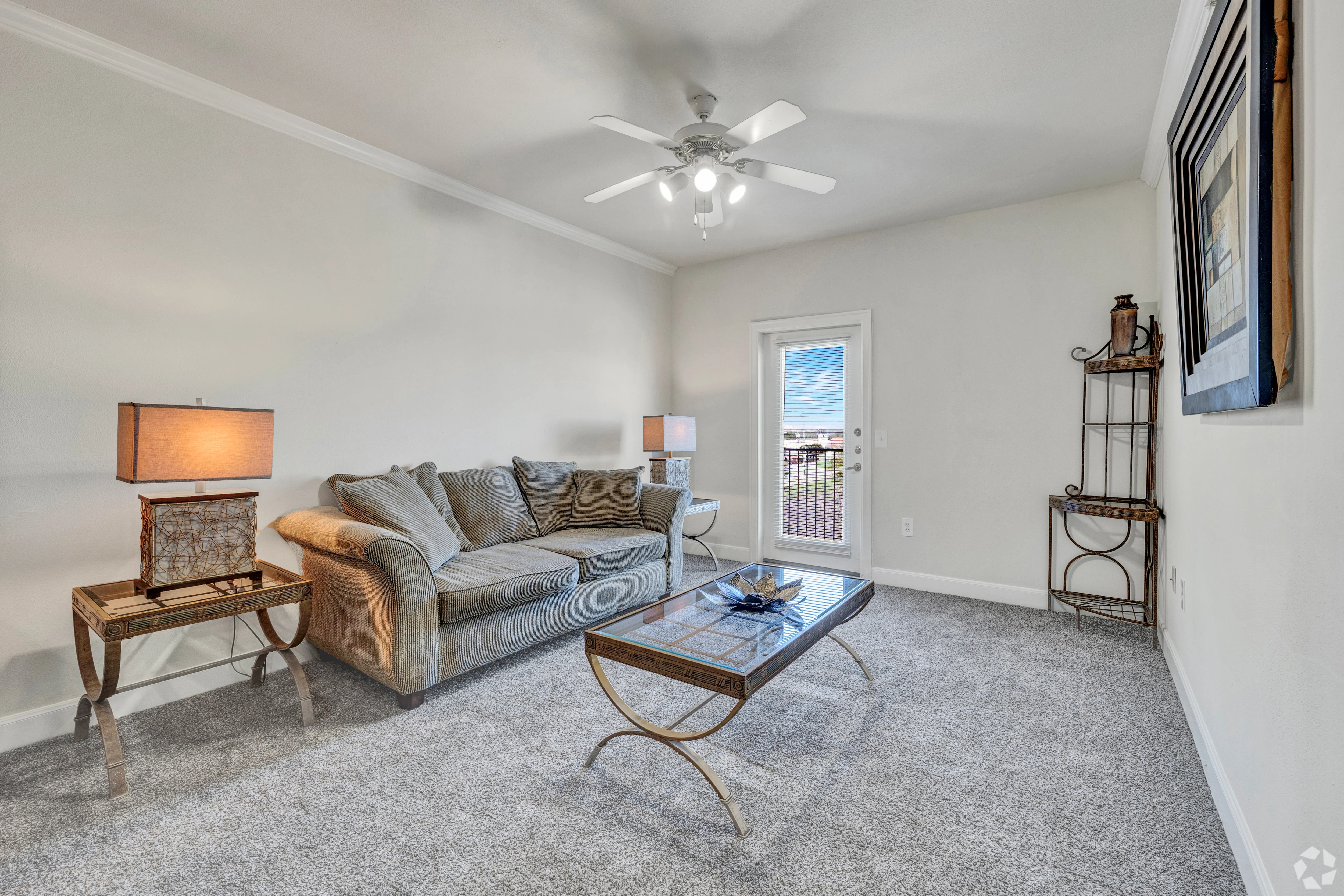 A cozy living room featuring a gray sofa, two lamps on side tables, and a glass coffee table. There's a door leading outside and light-colored walls with a ceiling fan.